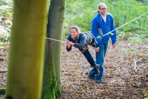 Frau schwingt an Seil zwischen Bäumen, Mann sichert sie am Boden
