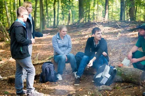 Gruppe diskutiert am Waldboden neben einem kleinen Feuer und Rucksäcken