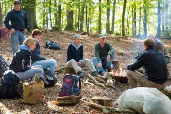 Gruppe sitzt um ein Lagerfeuer in einem Wald, Rucksäcke und Holz liegen herum
