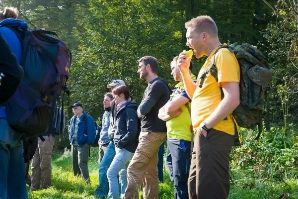 Gruppe von Personen mit Rucksäcken hört aufmerksam einer Erklärung im Wald zu