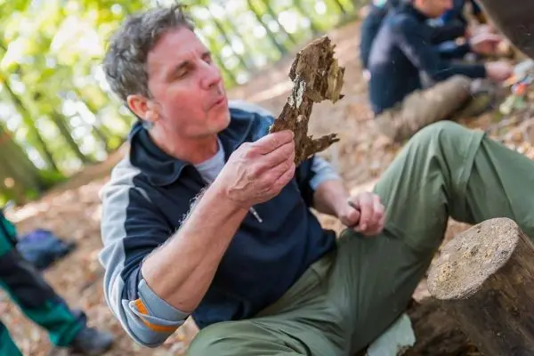 Holzstück wird mit einem Messer bearbeitet, während im Hintergrund weitere Personen arbeiten