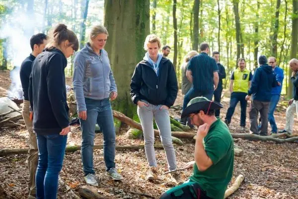 Mann demonstriert Holzarbeitstechniken auf dem Waldboden, umringt von Interessierten