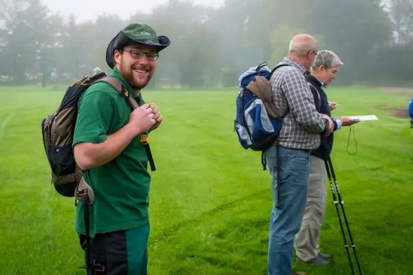 Mann mit Rucksack lächelt auf einer Wiese, während andere Notizen machen