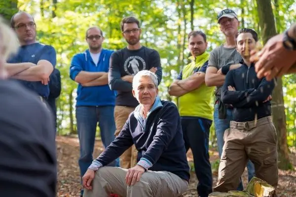 Mann sitzt auf einem Baumstamm, umgeben von mehreren Personen im Wald