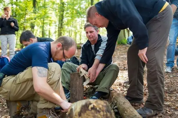 Zwei Personen bearbeiten Holz mit einem Messer, während andere zuschauen