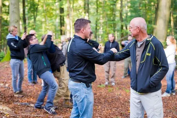 Zwei Personen üben eine Handgelenkstechnik im Wald, während andere zuschauen