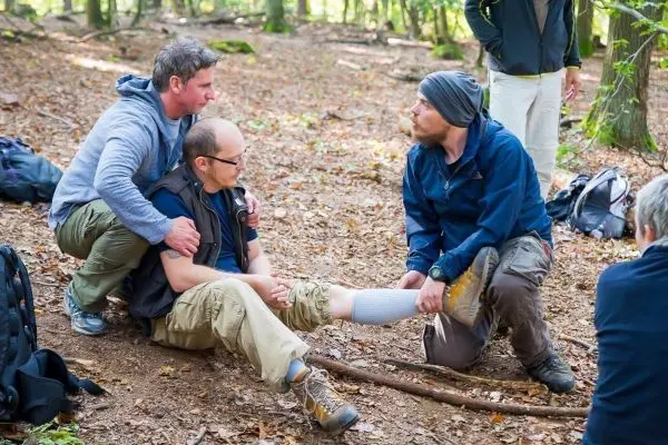 Zwei Personen unterstützen einen Verletzten im Wald, während ein Dritter die Verletzung untersucht