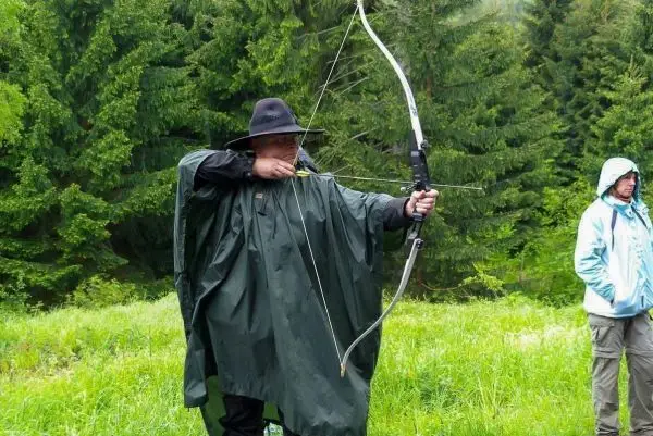 Bogenschütze spannt Pfeil in einem grünen Poncho an einem Waldrand