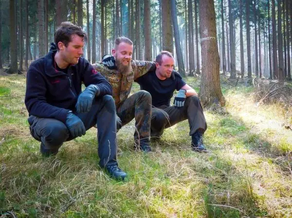Drei Personen hocken im Wald auf dem Boden und scheinen sich zu unterhalten