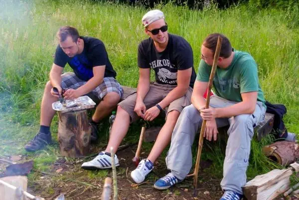 Drei Personen sitzen auf Holzstämmen und halten Stöcke in der Hand
