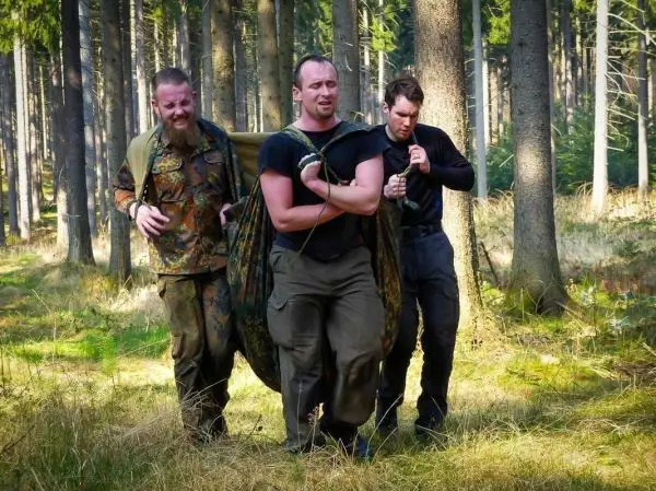 Drei Personen tragen eine improvisierte Trage durch einen Wald