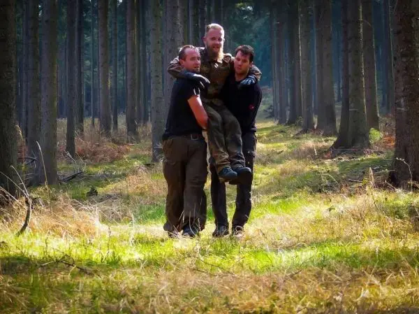 Drei Personen tragen einen Mann durch einen Wald auf einem grasbewachsenen Weg