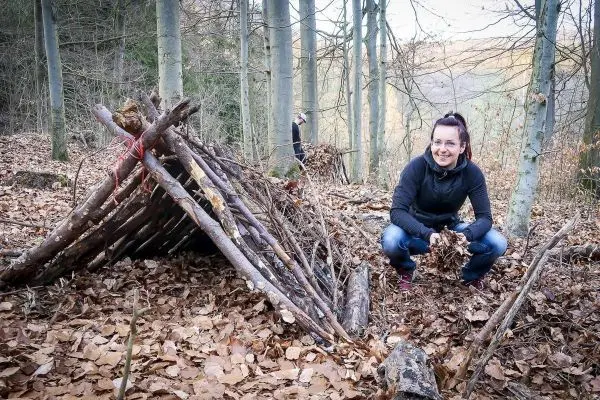 Frau platziert Laub vor einer selbstgebauten Tarp-Notunterkunft im Wald