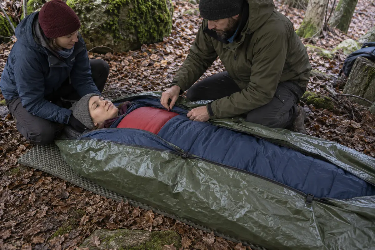 Unterkühlte Person liegt waagerecht auf einer Isomatte in einem Schlafsack mit wasserdichter Außenhülle und breiter Wärmequelle auf der Brust