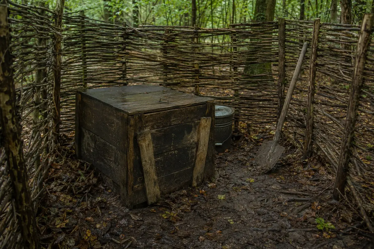 Verwitterte geschlossene Feldlatrine mit Flechtwand, Schaufel und Abdeckmaterial im Wald