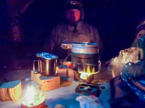 Hobo-Kocher mit Kessel auf einem Tisch, daneben brennendes Feuer und Geschirr