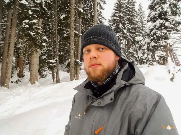 Mann in warmer Jacke steht im verschneiten Wald zwischen hohen Bäumen