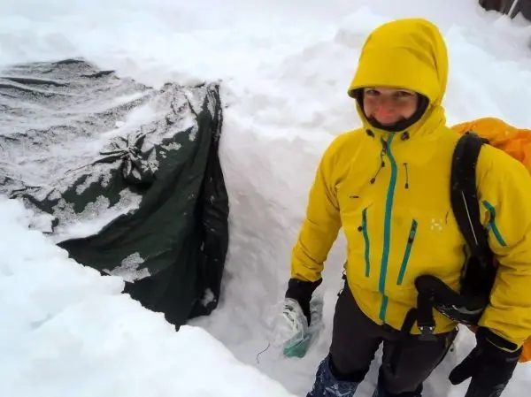 Person in gelber Jacke steht neben einer Tarp-Notunterkunft im Schnee