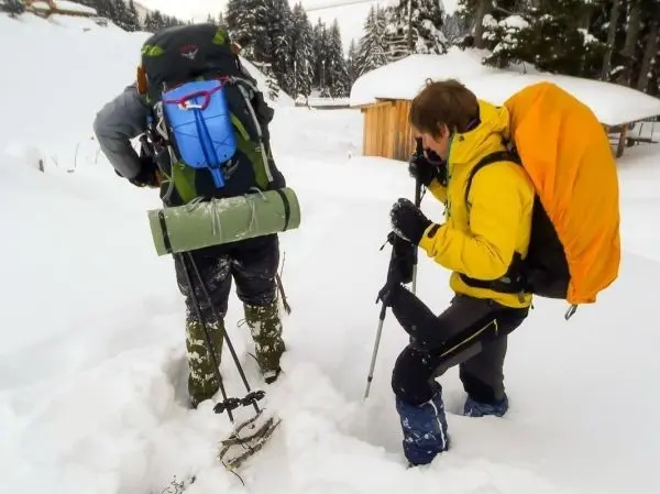 Person mit Rucksack untersucht Schnee, andere Person hält Skistöcke