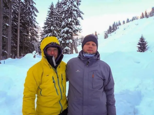 Zwei Personen in winterlicher Kleidung stehen im Schnee vor einer bewaldeten Landschaft