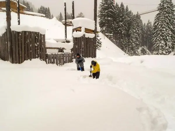 Zwei Personen schaufeln Schnee vor einer Holzkonstruktion im Winter